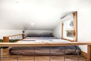 A bedroom with a sloped ceiling, a window with a view, a bed with striped bedding, and a wooden railing. - Impressive Intimate Tiny Cabin with Lofted Interior and Outdoor Shower in Texas (Wimberley)