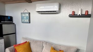 A living room with a sofa, pillows, a wall-mounted air conditioner, a microwave, and a refrigerator. - Off-Grid, Lakeside Tiny Home in Punta Gorda, Florida, Perfect for a Weekend Getaway (Punta Gorda)