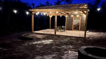 A covered outdoor seating area with a wooden structure, a fire pit, and string lights.