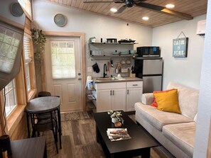 A compact kitchen with a microwave, refrigerator, and hanging utensils. There is a dining area with a table and chairs, and a sofa with a cushion.