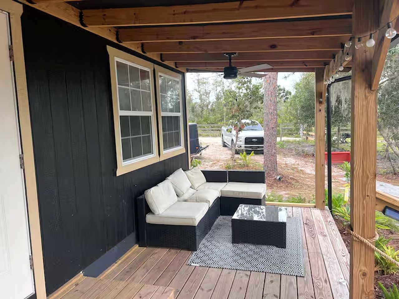 Un espacio de estar techado al aire libre con un sofá, una mesa de centro, una terraza de madera y un vehículo al fondo.