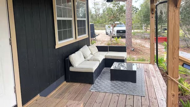 A covered outdoor seating area with a sofa and a coffee table, a wooden deck, and a view of a vehicle in the background. - Off-Grid, Lakeside Tiny Home in Punta Gorda, Florida, Perfect for a Weekend Getaway (Punta Gorda)