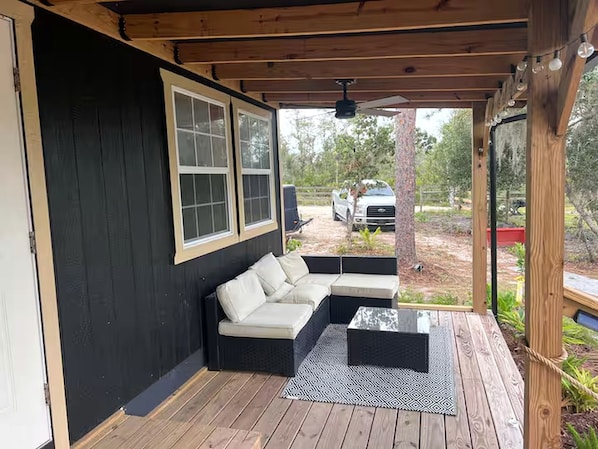 A covered outdoor seating area with a sofa and a coffee table, a wooden deck, and a view of a vehicle in the background.