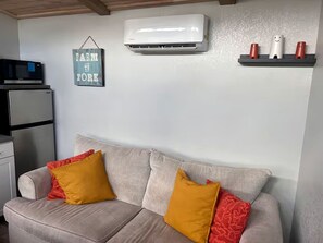 A living room with a sofa, pillows, a wall-mounted air conditioner, a microwave, and a refrigerator.
