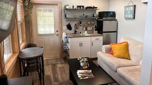 A compact kitchen with a microwave, refrigerator, and hanging utensils. There is a dining area with a table and chairs, and a sofa with a cushion. - Off-Grid, Lakeside Tiny Home in Punta Gorda, Florida, Perfect for a Weekend Getaway (Punta Gorda)