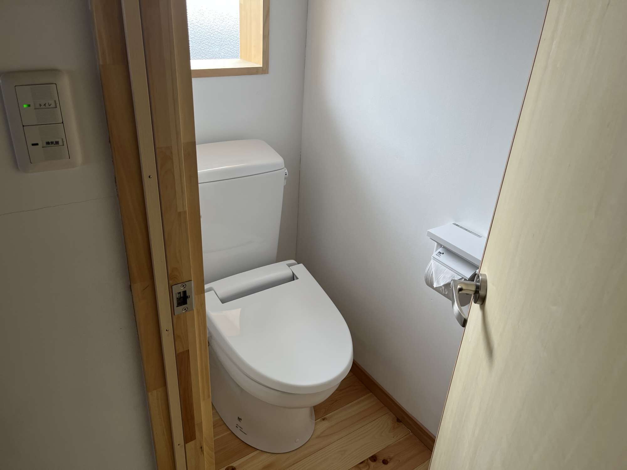 A small, clean bathroom with a toilet, a wooden floor, and a window above the toilet.
