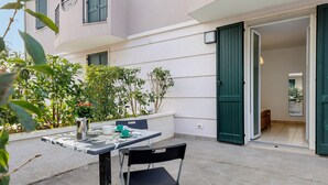 Une terrasse extérieure avec une table et des chaises, un pot de fleurs et l’entrée d’un bâtiment.