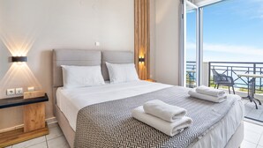 Habitación de hotel moderna con una cama grande, una mesita de noche con lámpara, un balcón con mesa y sillas, y vista al cielo.
