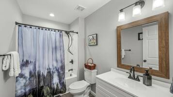Une salle de bain avec un rideau de douche représentant une scène de forêt, des toilettes, un lavabo avec un miroir et un panier fixé au mur.