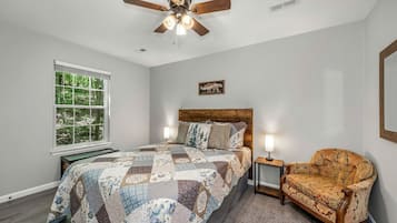 A bedroom with a wooden bed, a quilt, a chair, a nightstand, a window with a view of trees, and a ceiling fan.