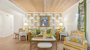 Una sala de estar con un sofá verde, un sillón amarillo y una mesa de centro con libros. Hay dos lámparas sobre mesitas auxiliares y un cuadro en la pared.