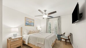 Un dormitorio con cama, mesitas de noche, una silla, ventilador de techo, televisor y un cuadro en la pared.