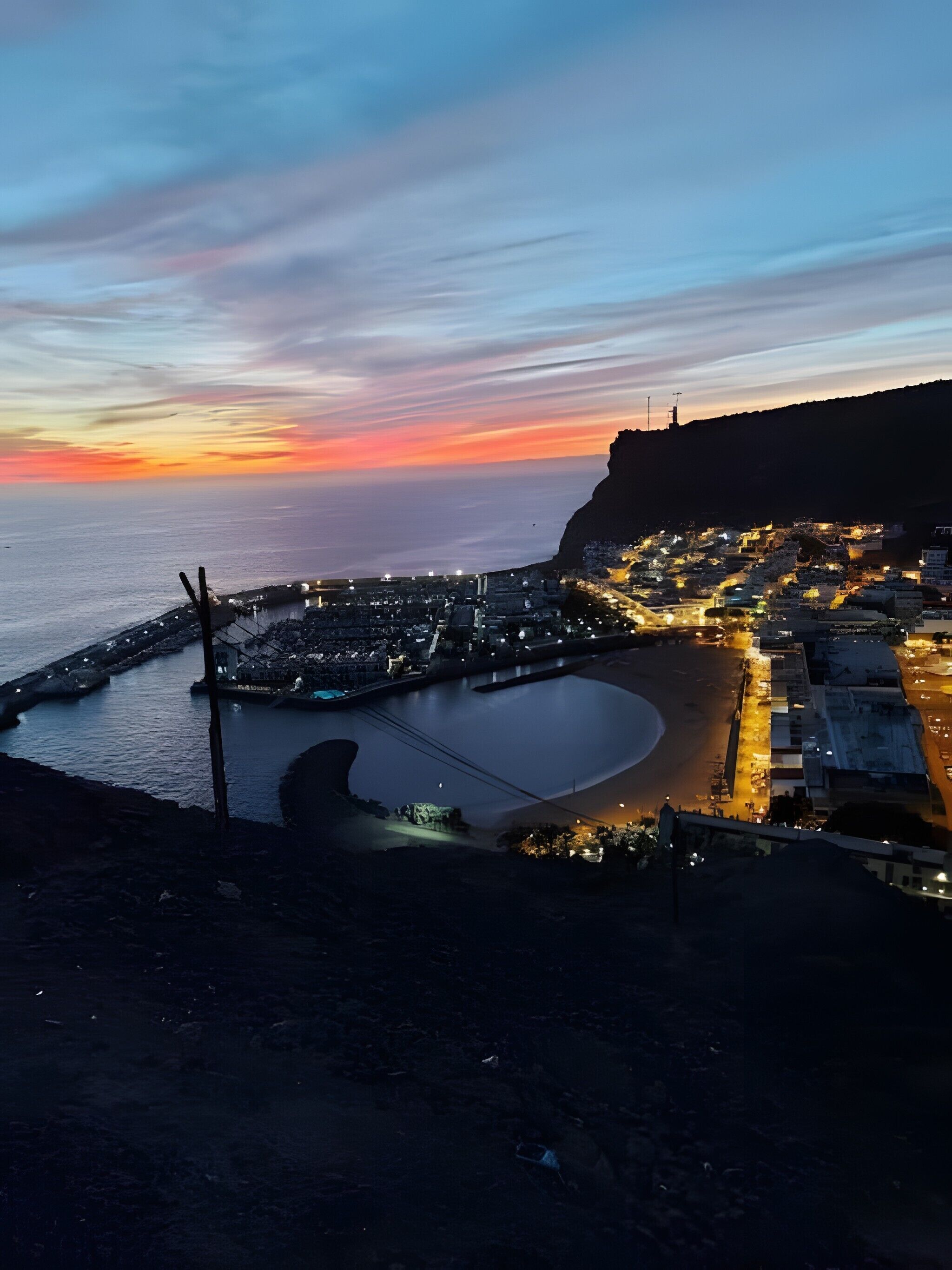 A coastal town at dusk with a prominent cliff, a marina, and illuminated buildings.