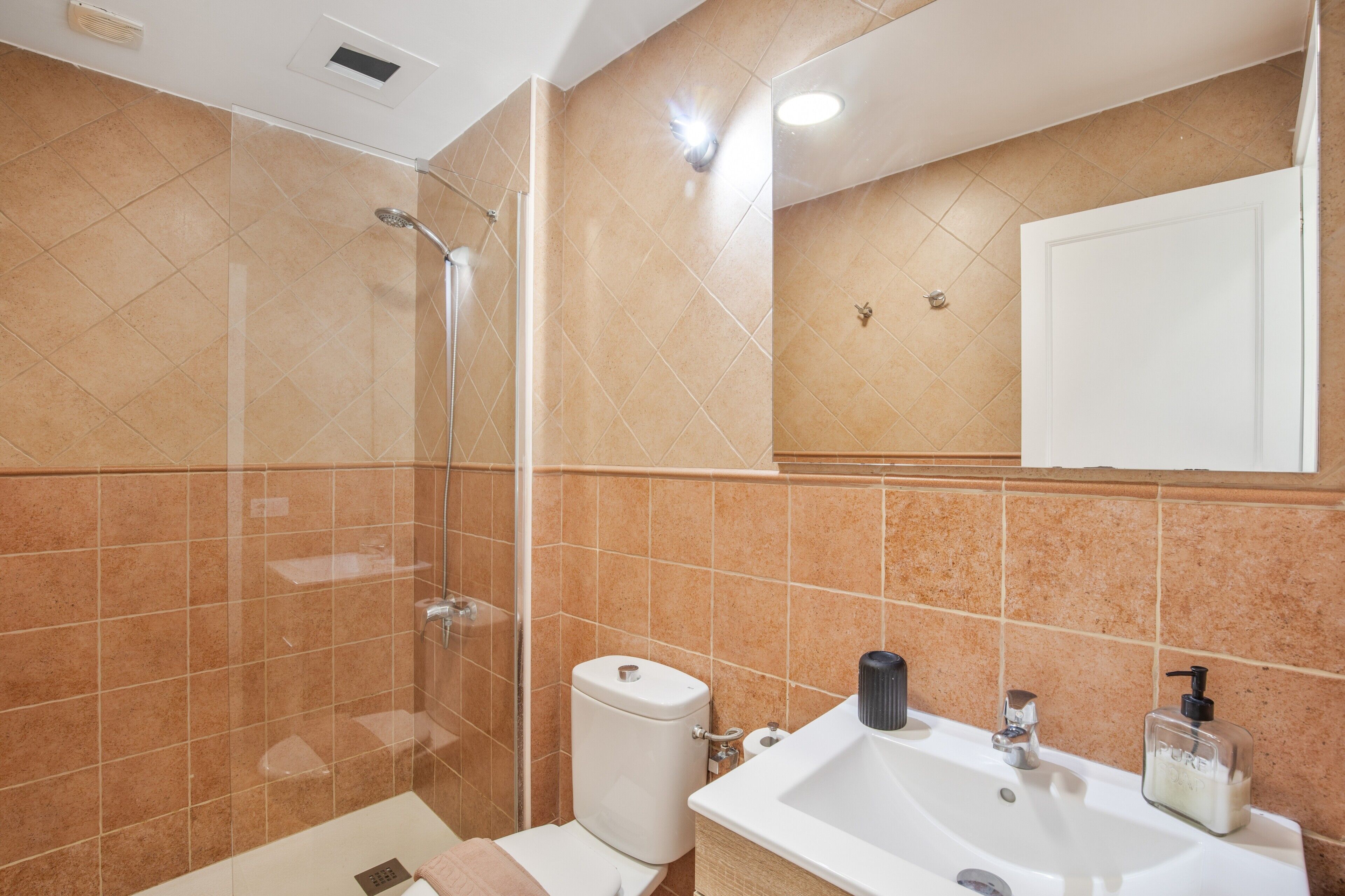 Un baño con cabina de ducha de vidrio, inodoro, lavamanos con dispensador de jabón y espejo sobre el lavamanos.