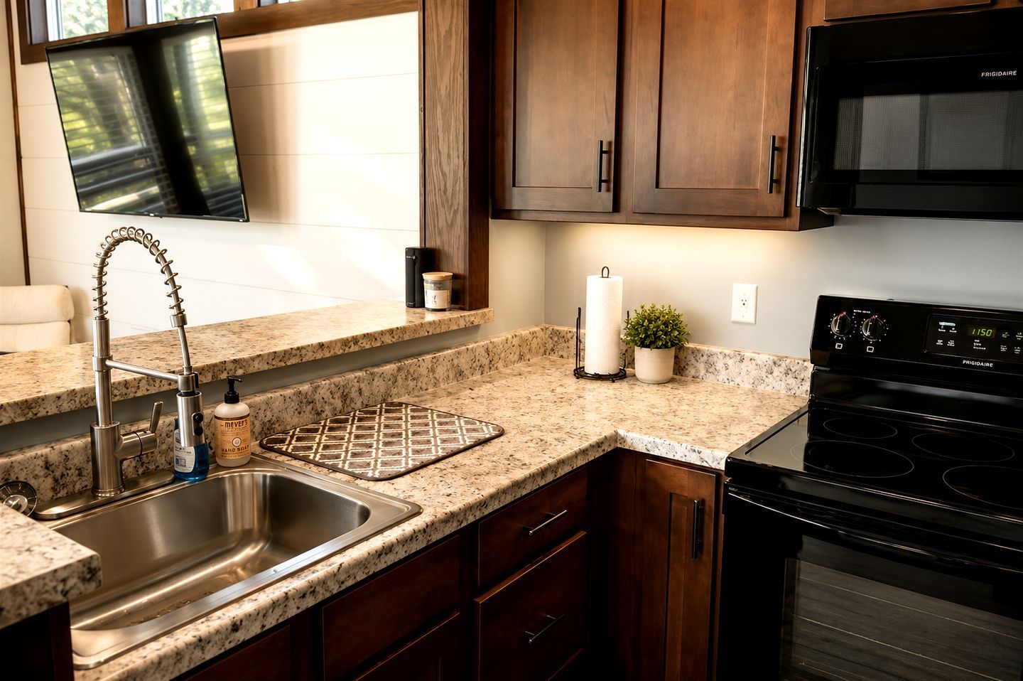 A kitchen with granite countertops, a stainless steel sink, a black stove, wooden cabinets, and a flat-screen TV mounted above the counter.