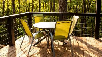 A wooden deck with a table and chairs, surrounded by a metal railing, overlooking a forest.