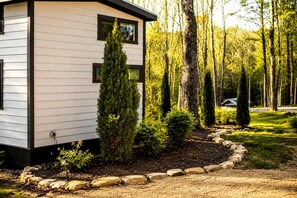 A modern cabin with a white exterior and black trim, surrounded by a landscaped garden with various shrubs and a stone border.