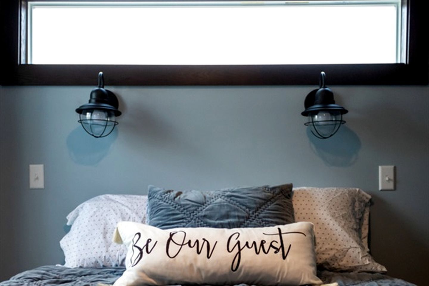 Une chambre à coucher avec un lit, deux lampes fixées au mur et un coussin orné de la phrase « Soyez nos invités ».
