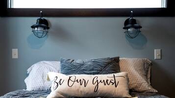 Une chambre à coucher avec un lit, deux lampes fixées au mur et un coussin orné de la phrase « Soyez nos invités ».