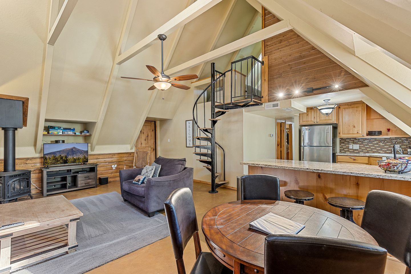 A loft-style living space with a high ceiling, a wooden staircase, a kitchen area, and a seating area with a table and chairs.