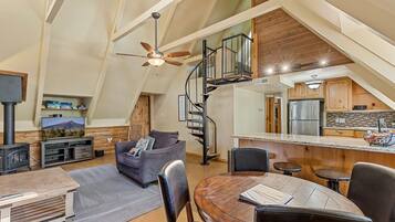 A loft-style living space with a high ceiling, a wooden staircase, a kitchen area, and a seating area with a table and chairs.