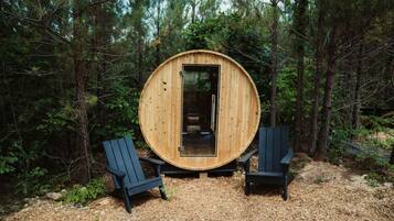 A wooden sauna in a forest with two Adirondack chairs.