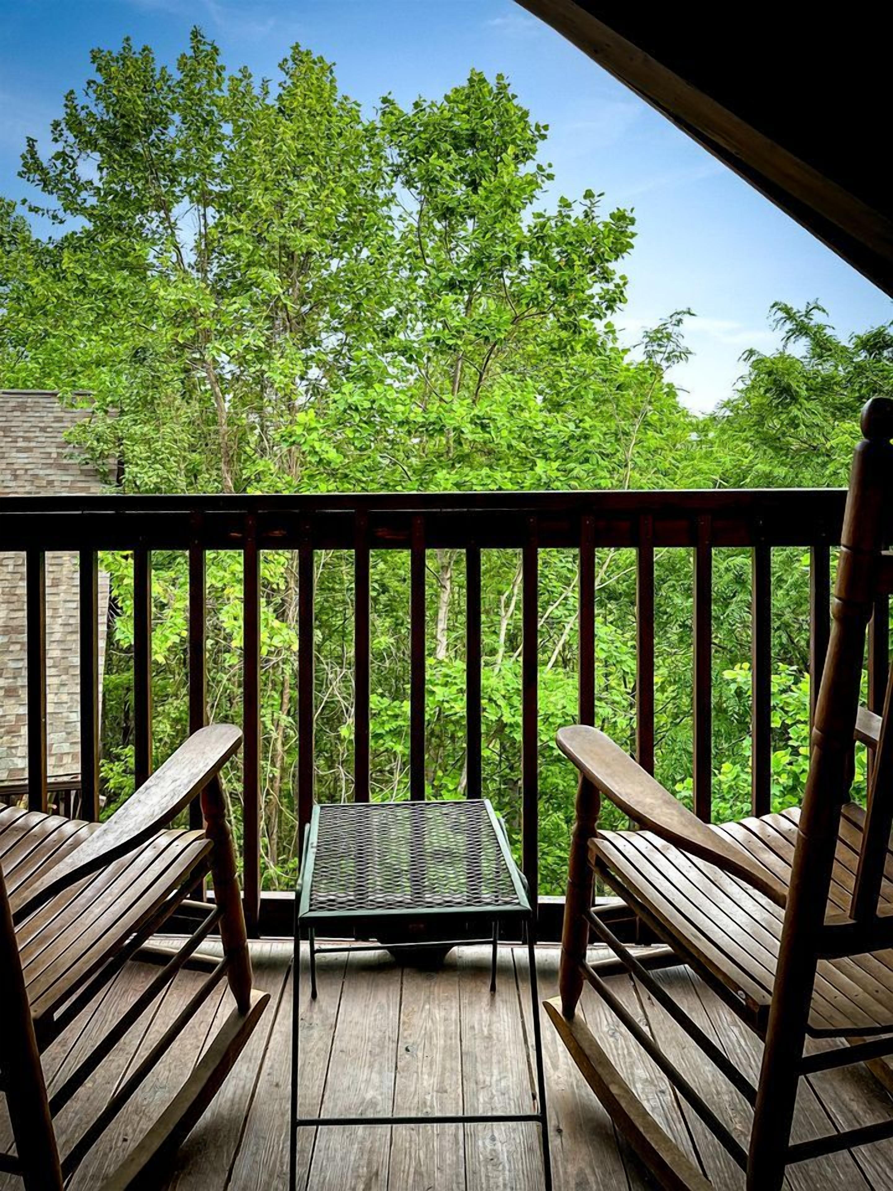 Une terrasse en bois avec deux chaises et une petite table, entourée d’arbres verdoyants.