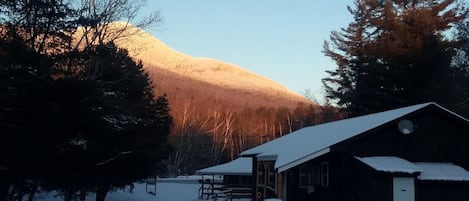 Una cabaña con techo nevado, un porche de madera y una montaña al fondo.