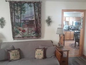 Ein Wohnzimmer mit Sofa, Tisch mit Lampe, Wandteppich und Blick in eine Küche.