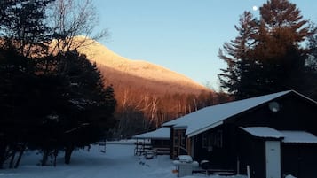 Una cabaña con techo nevado, un porche de madera y una montaña al fondo.