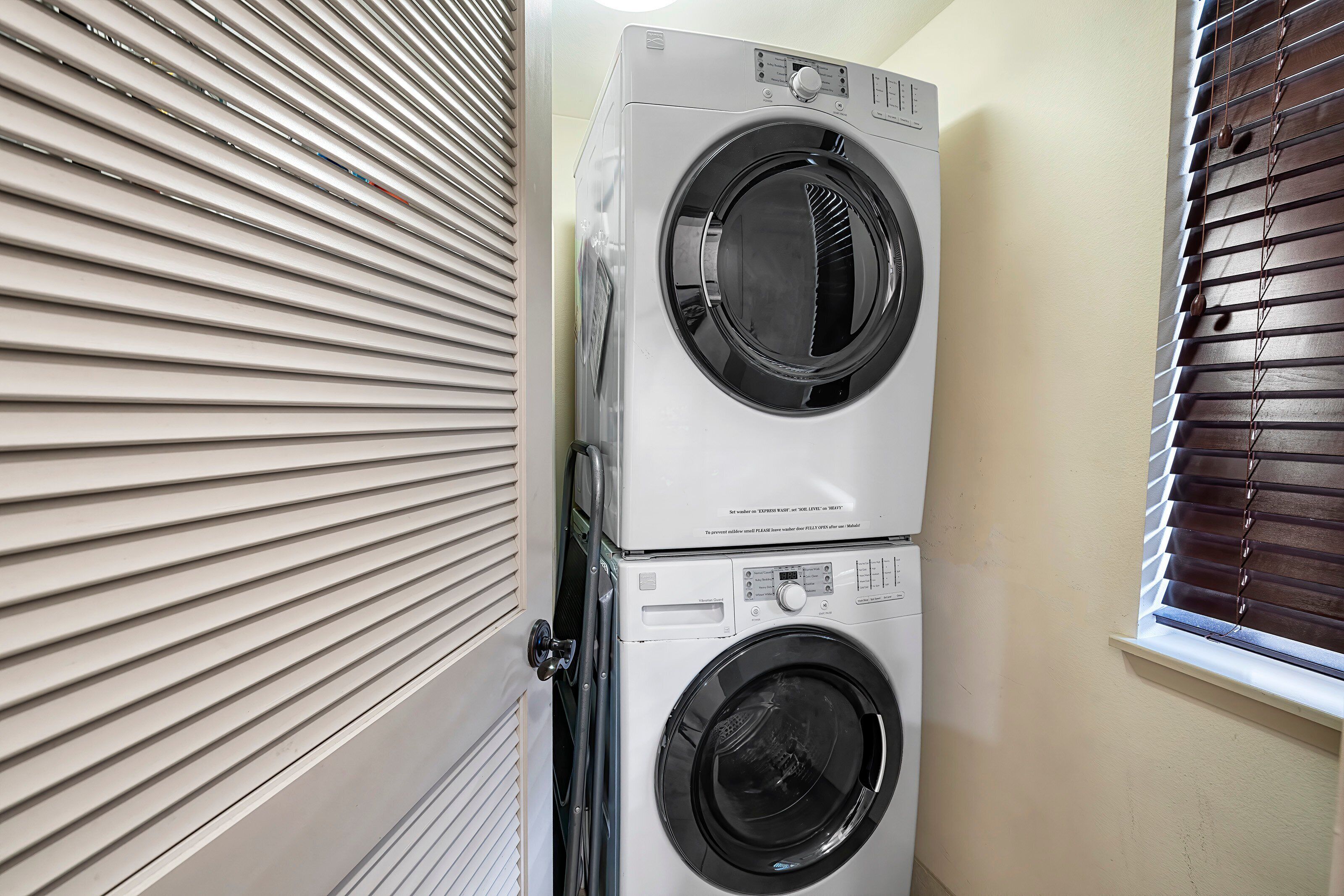 A stacked washer and dryer unit in a laundry room with blinds on the windows.