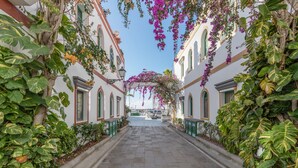 A narrow street flanked by buildings with large windows and green shutters, leading to a white building with a visible entrance. - Vacation Studio 'Vv Ancla' with Mountain View, Wi-Fi and Air Conditioning (Puerto de Mogán)