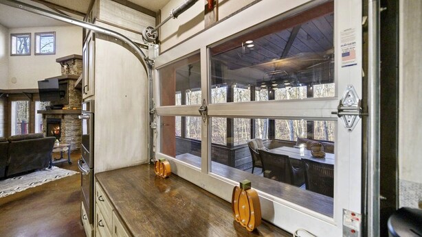 Um quarto com lareira, uma área de jantar com mesa e cadeiras, e uma janela grande com vista para árvores.