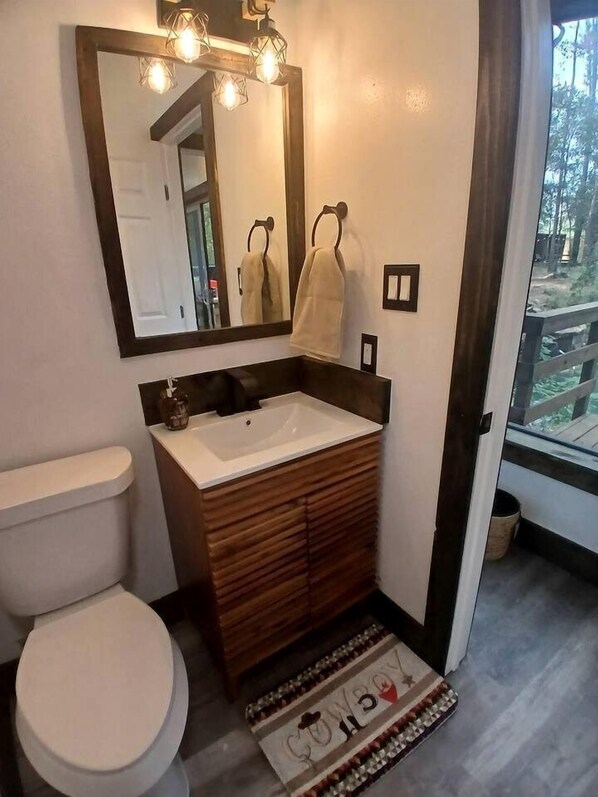 A bathroom with a white toilet, wooden vanity, a large mirror with lights, and a view of a deck through a glass door.