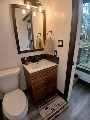Une salle de bain avec des toilettes blanches, un meuble-lavabo en bois, un grand miroir avec éclairage intégré et une vue sur une terrasse accessible par une porte vitrée.
