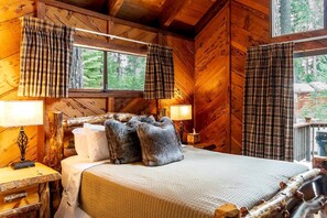 Une chambre à coucher dans un chalet en bois, avec un lit, deux oreillers, une table de chevet, une lampe et des rideaux à carreaux.