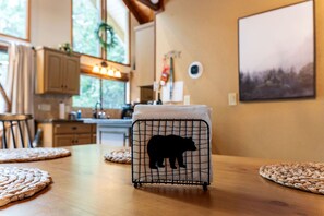 Une cuisine avec des meubles de rangement en bois, une fenêtre décorée d’une guirlande, une table avec des sets de table en fibres tressées et un panier en fil métallique orné de la silhouette d’un ours.