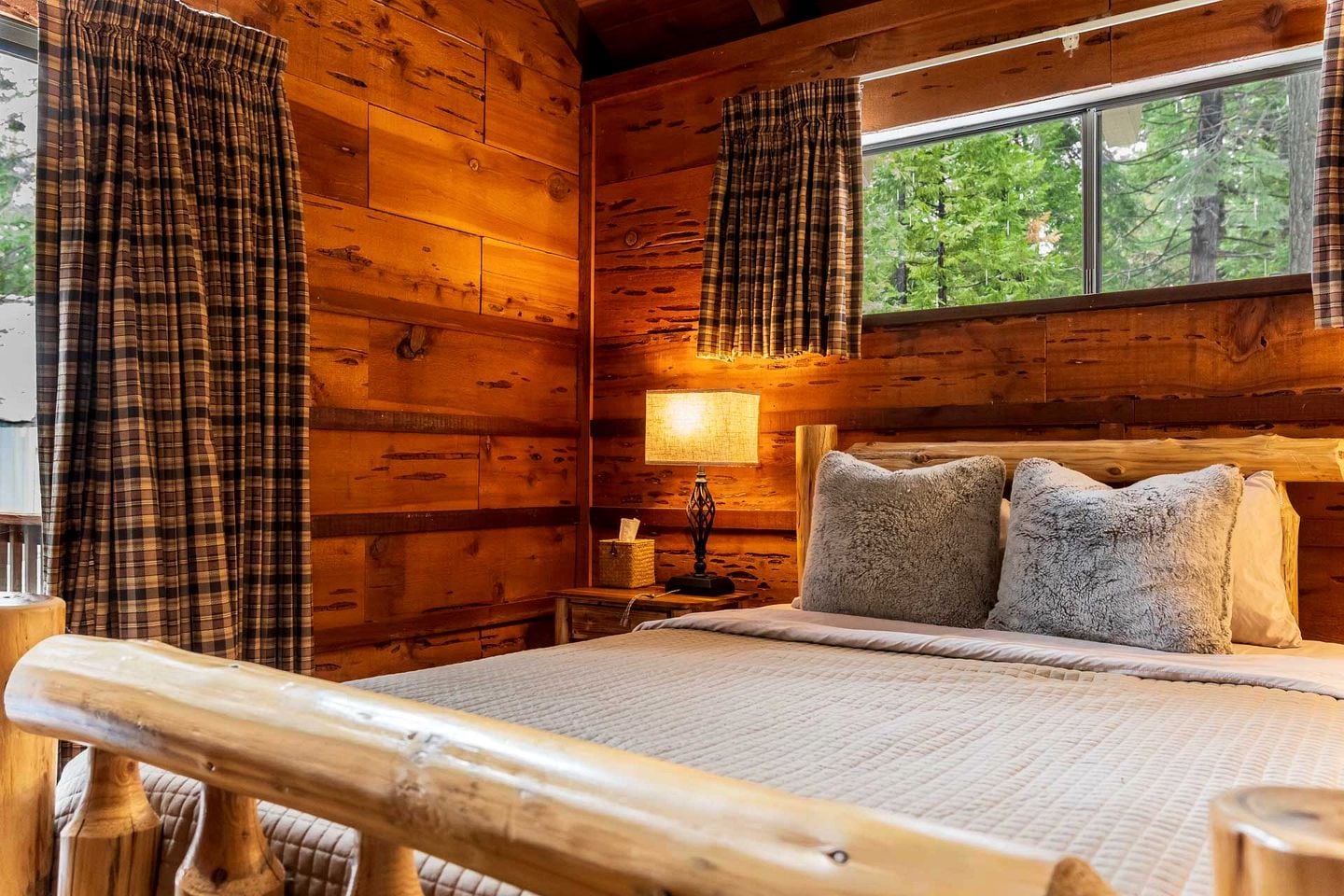 A log cabin bedroom with a bed, plaid curtains, a window with a view of trees, and a wooden headboard.