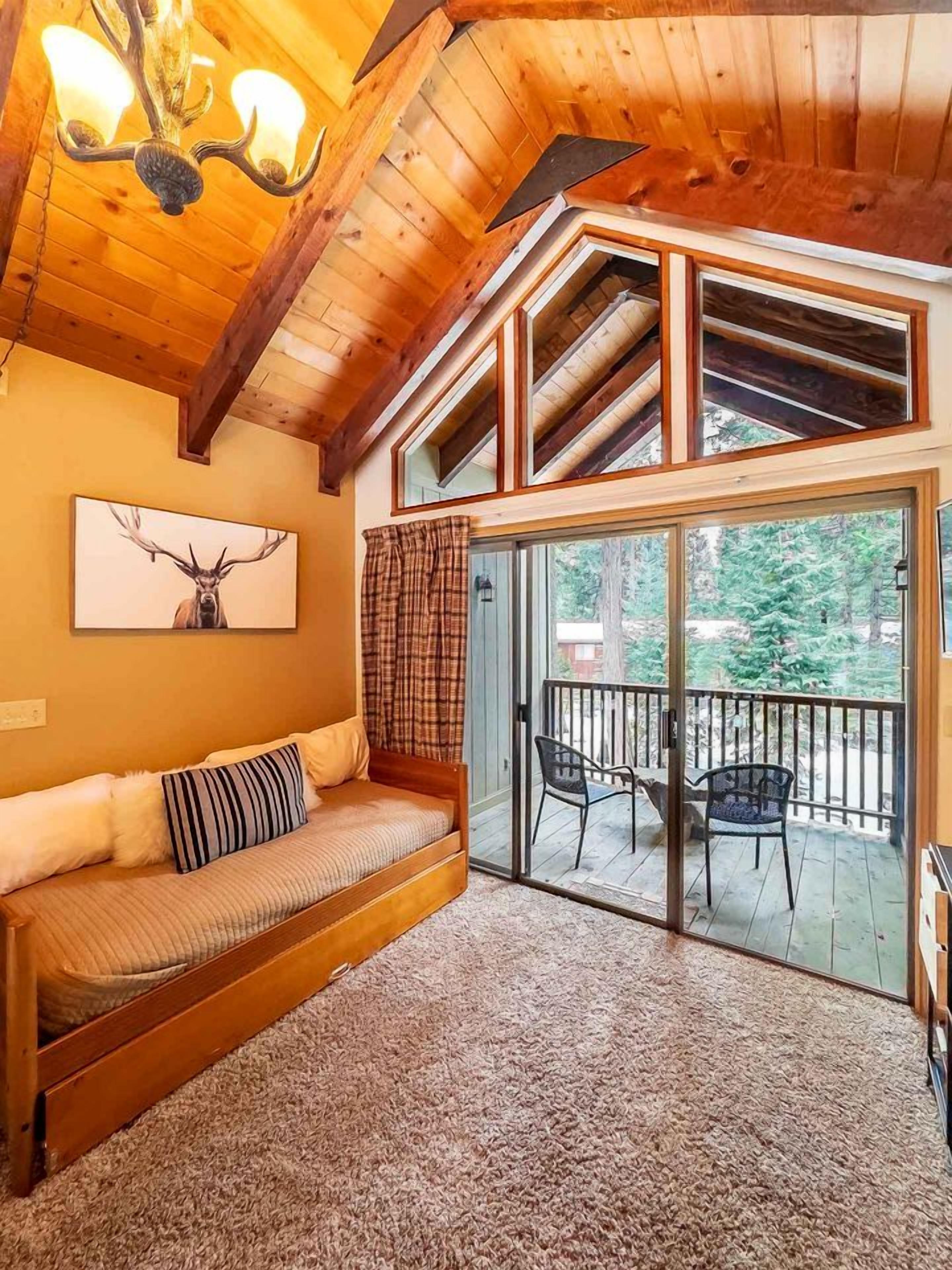 A cozy cabin interior with a wooden ceiling, a sofa with cushions, a large window, and a view of the outdoors.