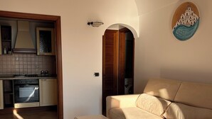 A living room with a beige sofa and a white table, a kitchen with a stove and cabinets, and a decorative wall piece.