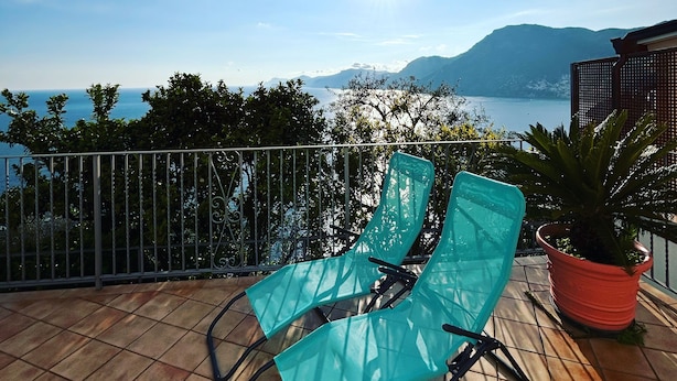 A terrace with two turquoise chairs and a potted plant, overlooking a lake and mountains.
