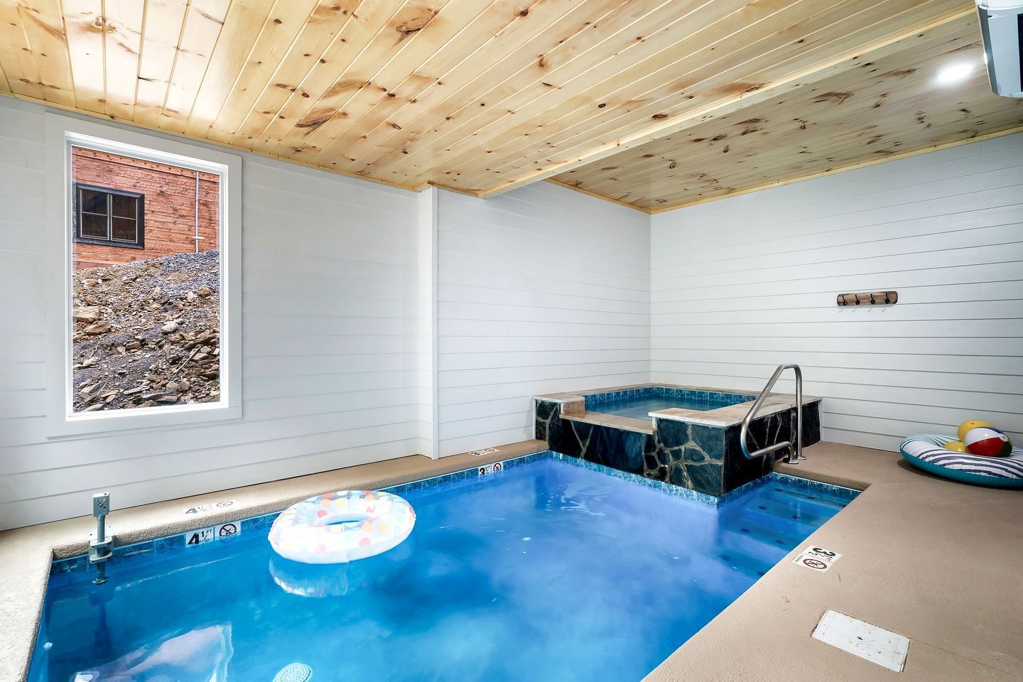 A swimming pool with a hot tub, wooden ceiling, and a window overlooking a rocky landscape.