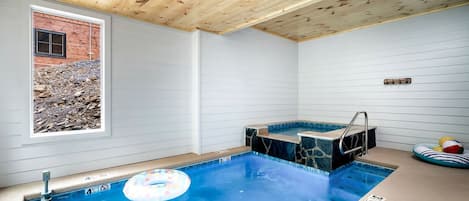 A swimming pool with a hot tub, wooden ceiling, and a window overlooking a rocky landscape.