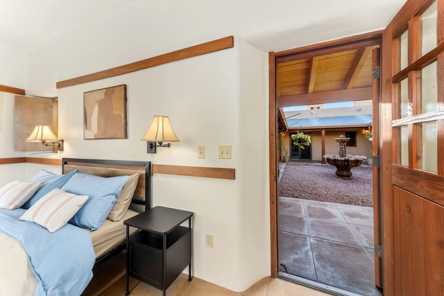 A bedroom with a bed, bedside tables, wall-mounted lamps, and a view of an outdoor courtyard with a fountain.
