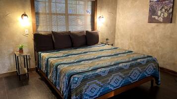Un dormitorio con una cama, dos luces fijadas a la pared, un ventanal con persianas, una mesita con una planta y un cuadro en la pared.