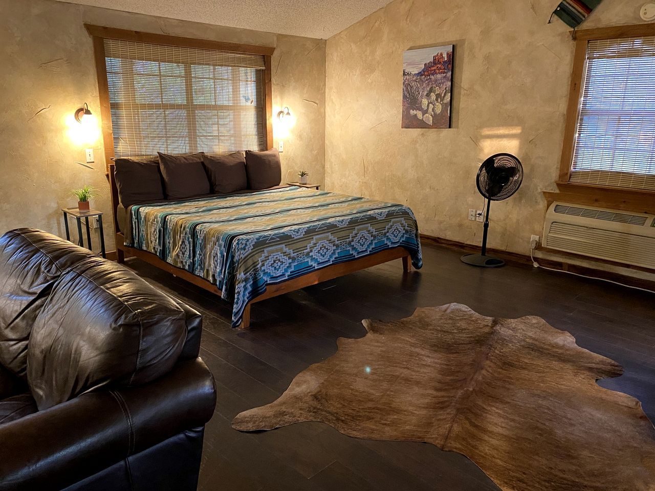 A bedroom with a bed, a leather sofa, a rug, a fan, and a painting on the wall.