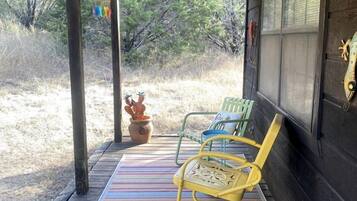 A wooden deck with a striped mat, two chairs, a potted plant, and a window with a lizard decoration.