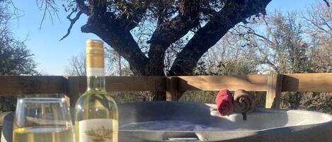 A picnic setup with a bottle of wine, a glass, and a plate of snacks on a table, with a hot tub and a tree in the background.
