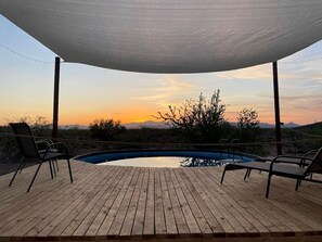 A wooden deck with a pool, lounge chairs, and a view of the sunset.