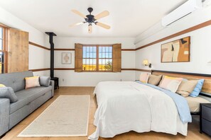 A bedroom with a bed, a sofa, a window with a view, a ceiling fan, and an air conditioning unit.